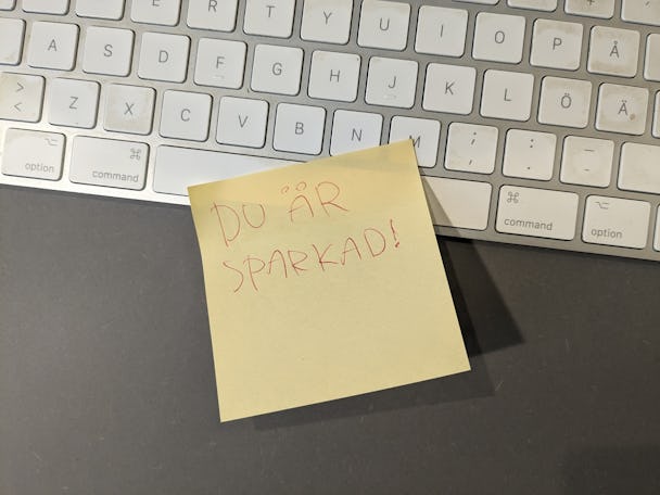 En klisterlapp med frasen "du är sparkad!" handskriven på den, placerad på en grå yta bredvid ett vitt datortangentbord.