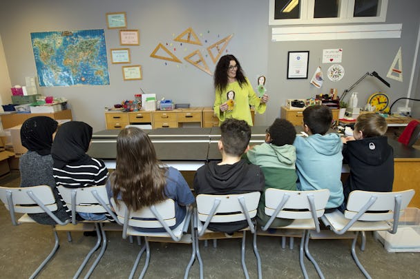 En grupp barn sitter vid ett bord i ett klassrum. Lärarassistent Lena Rilling håller upp två teckningar för gruppen.