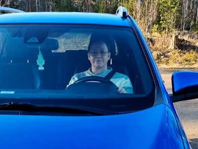 En person som sitter i förarsätet i en blå bil parkerad vid sidan av en väg med träd i bakgrunden.