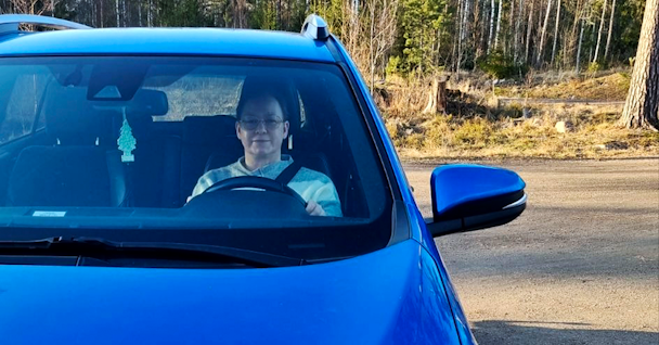 En person som sitter i förarsätet i en blå bil parkerad vid sidan av en väg med träd i bakgrunden.
