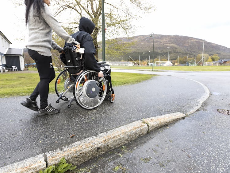 Kvinna som hjälper en person i rullstol på en asfalterad stig utomhus.