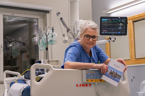 En vårdpersonal i ett sjukhusrum som håller en dagbok, med medicinska monitorer och utrustning i bakgrunden.