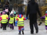 En grupp små barn som bär synliga västar som håller hand och går med en vuxen på en stadstrottoar.