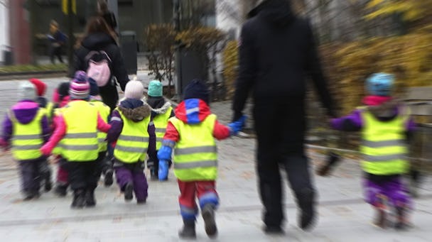 En grupp små barn som bär synliga västar som håller hand och går med en vuxen på en stadstrottoar.
