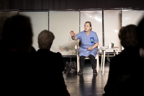 En kvinna i scrubs som sitter och talar under en presentation eller ett möte, med en publik vänd mot henne.