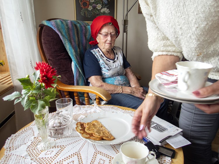 En äldre kvinna som sitter vid ett bord tittar på när någon serverar henne en kopp te och en tallrik med mat.