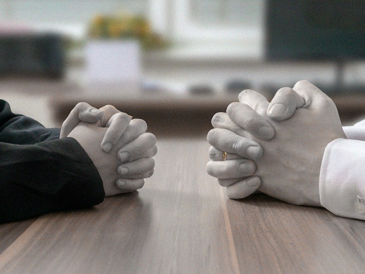 Två individer som sitter mittemot varandra, med händerna sammanslagna på ett bord, i en gest som antyder en seriös konversation eller förhandling.