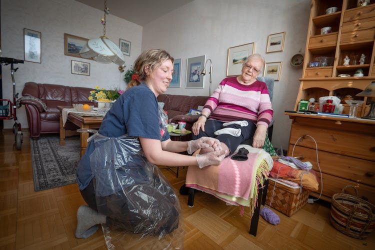 Hemvårdare som ger fotvård till en äldre kvinna i vardagsrumsmiljö.