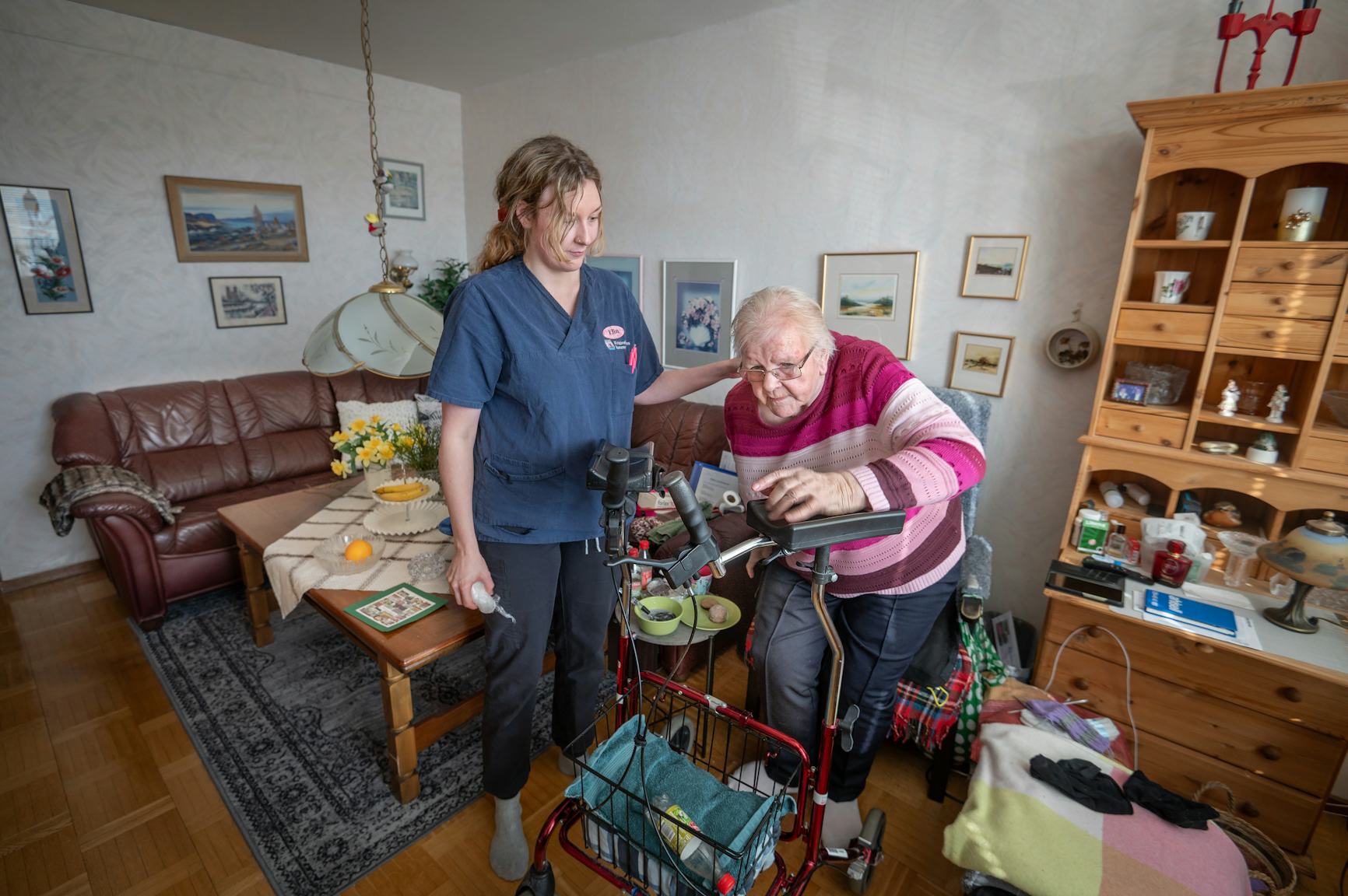 En vårdpersonal hjälper en äldre dam med hjälp av en rollator i ett mysigt vardagsrum.