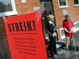 Fokusera på en röd "strejk!" skylt, med out-of-fokus arbetare som pratar i bakgrunden nära en byggnad.
