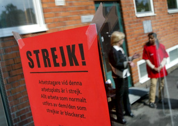 Fokusera på en röd "strejk!" skylt, med out-of-fokus arbetare som pratar i bakgrunden nära en byggnad.