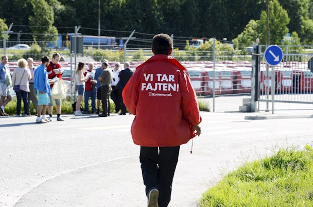 Person i röd jacka med texten "vi tar fajten!" på ryggen går ifrån en samlad grupp människor vid en parkering.