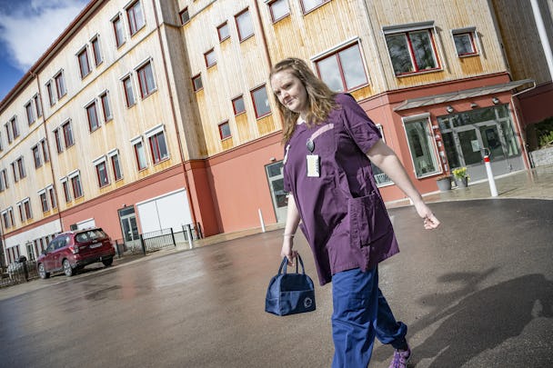 En vårdpersonal i sjukhuskläder promenerar utanför en modern sjukhusbyggnad samtidigt som hon bär på en väska och ler.