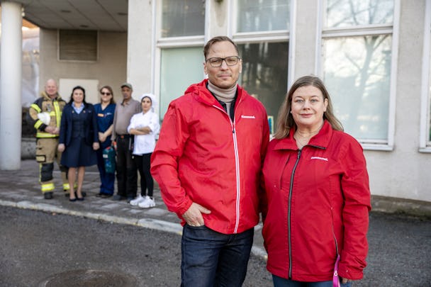 Två personer i röda jackor står framför en grupp nödpersonal och åskådare utanför en byggnad.