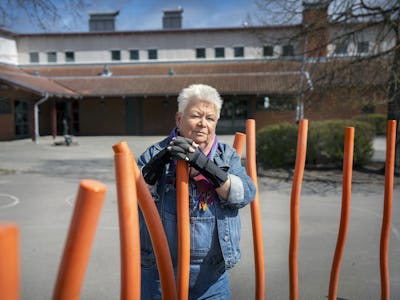 Äldre kvinna med kort vitt hår i jeansjacka och halsduk som står bakom orange stolpar på en innergård.