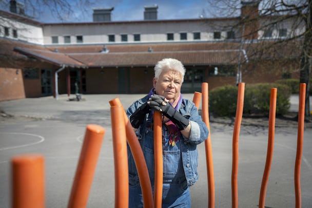 Äldre kvinna med kort vitt hår i jeansjacka och halsduk som står bakom orange stolpar på en innergård.
