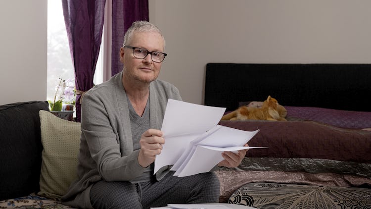 Medelålders man med glasögon och grå kofta på en soffa och granskar dokument, ingefära katt i bakgrunden.