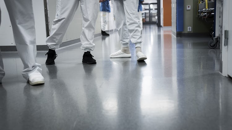 Alt text: Två sjukvårdspersonal som går längs en sjukhuskorridor, sedda från midjan och ner, en i svarta skor och den andra i vita.
