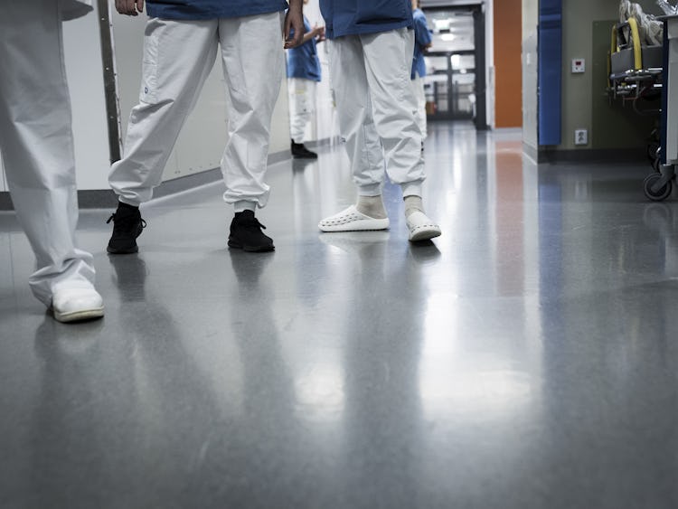 Alt text: Två sjukvårdspersonal som går längs en sjukhuskorridor, sedda från midjan och ner, en i svarta skor och den andra i vita.