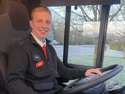 Buschaufför med uniform och namnskylt sitter bakom ratten i en buss.