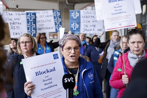 En grupp sjukvårdspersonal strejkar och håller protestskyltar. En kvinna i spetsen ses tydligt hålla i en skylt där det står "blockad".