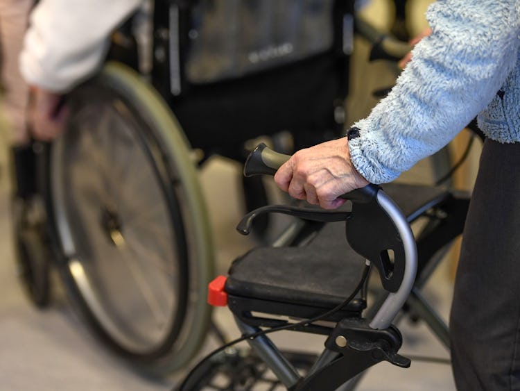 Ett förstorat foto på en äldre individs hand vilande på en rullatorshandtag, med en annan person i rullstol synligt i bakgrunden.