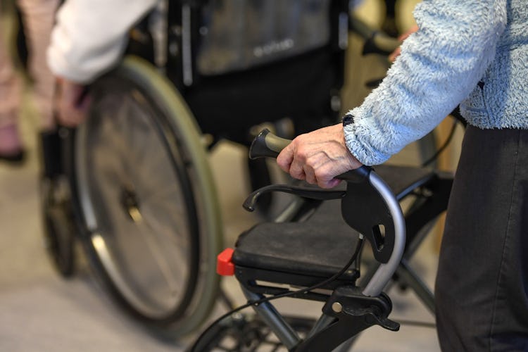 Ett förstorat foto på en äldre individs hand vilande på en rullatorshandtag, med en annan person i rullstol synligt i bakgrunden.
