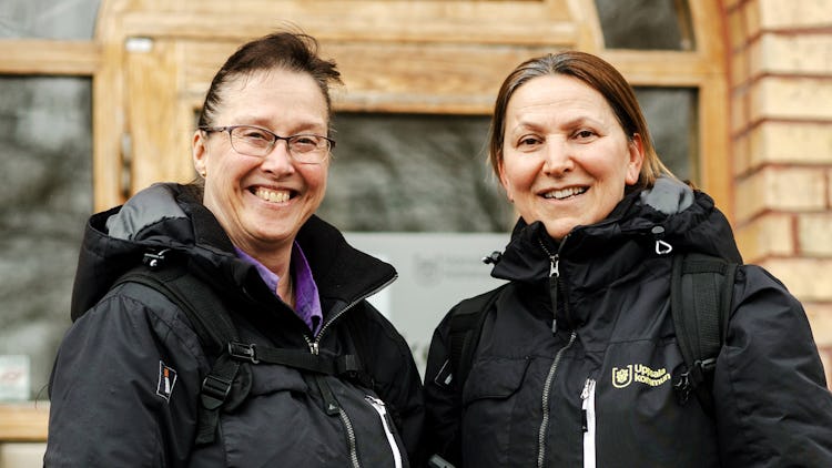 Två kvinnor i svarta jackor och ryggsäckar står framför en träbyggnad och ler mot kameran.