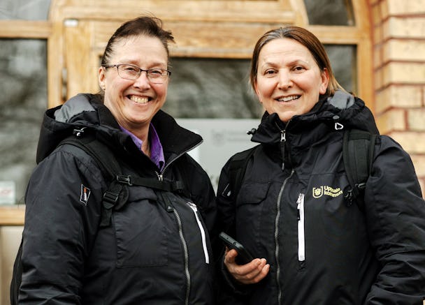 Två kvinnor i svarta jackor och ryggsäckar står framför en träbyggnad och ler mot kameran.