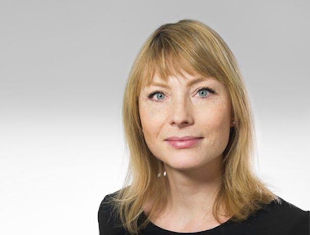 En professionell porträttbild av en kvinna med blont hår och lugg, blå ögon, bär diskret smink och små örhängen på en ljus bakgrund.