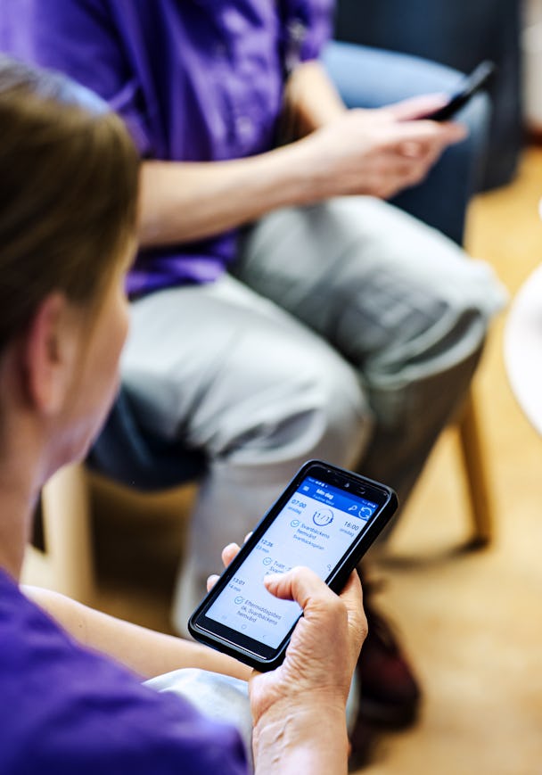 Två personer använder smartphones i ett väntrum, med fokus på en kvinnas händer som betraktar en hälsorelaterad app på sin enhet.