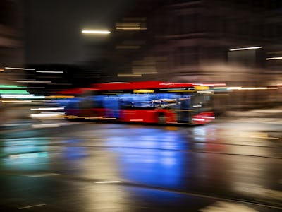 En oskarp rörelsebild av en röd buss som kör genom en regntung gata på nattetid, med ljusstrimmor och reflektioner.