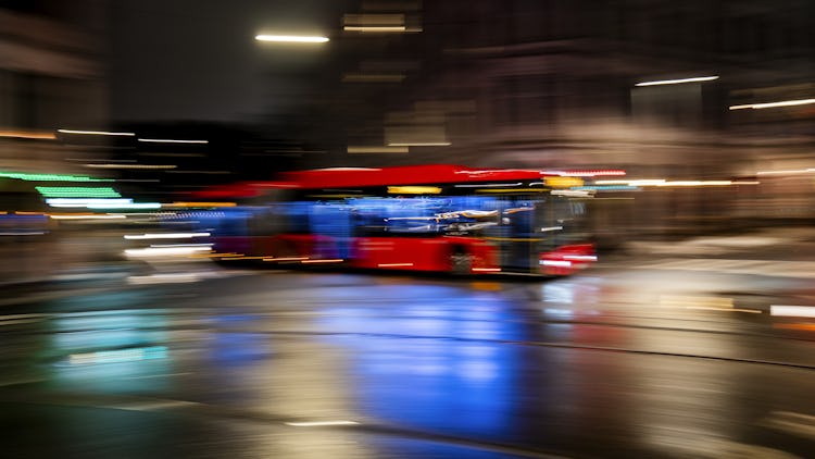 En oskarp rörelsebild av en röd buss som kör genom en regntung gata på nattetid, med ljusstrimmor och reflektioner.