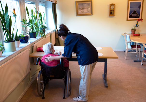 En vårdgivare i blå uniform hjälper en äldre kvinna i rullstol vid ett bord i ett upplyst rum med växter och bilder på väggarna.