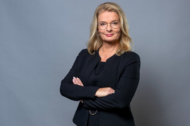Skolministern Lotta Edholm i svart kostym och glasögon står med korsade armar mot en grå bakgrund.