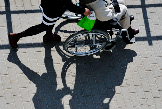 Två personer, en gående och en i rullstol, på en kullerstensgata med sina skuggor synliga på marken.