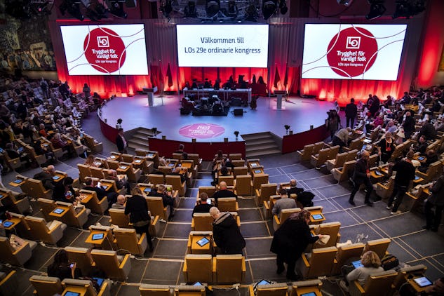 En konferenshall med sittande deltagare som umgås. Två stora skärmar visar texten "Välkommen till LOs 29:e ordinarie kongress" på svenska. En scen med ett band är uppställd i bakgrunden.