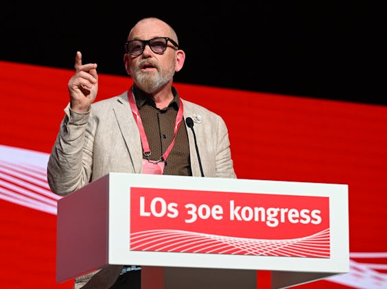 Johan Lindholm med glasögon och kavaj talar vid ett podium framför en röd bakgrund som visar texten "LOs 30e kongress" i vitt.