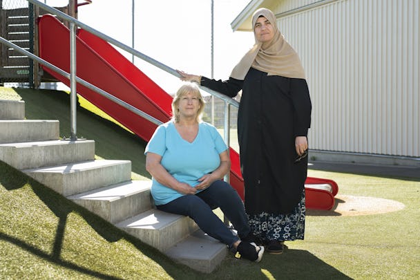 Två kvinnor, Helene Joabsson och Maida Haidare, en sittande och en stående, poserar på trappsteg vid en rutschkana utanför en förskola. Den sittande kvinnan har på sig en ljusblå skjorta och den stående kvinnan bär huvudduk och lång kappa.