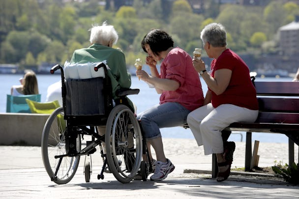 Tre personer äter glass utomhus; en av dem sitter i rullstol medan de andra två sitter på en bänk. I bakgrunden syns en vattensamling och ett grönt landskap.