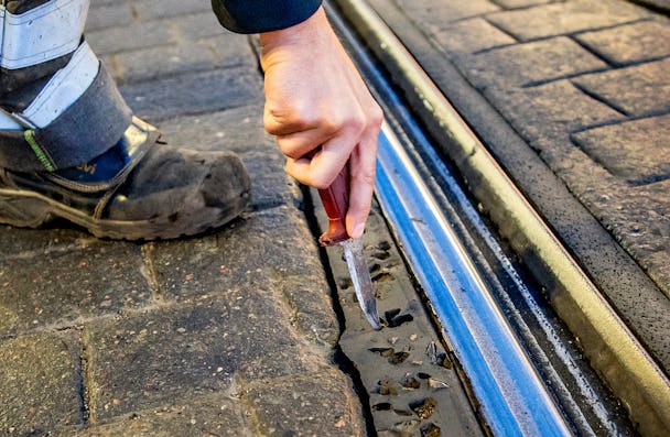 En person i arbetsstövlar använder ett kniv för att rensa bort skräp från en spårvägsräls som är inbäddad i en stenbelagd yta.