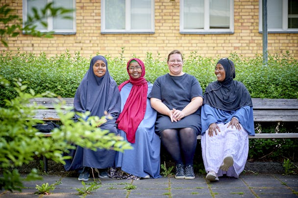 Fyra kvinnor sitter på en bänk utanför en byggnad av tegel och ler. Tre av dem bär hijab och färgglada kläder, medan den fjärde har på sig en svart klänning. Gröna växter omger bänken.