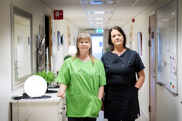 Två kvinnor i sjukvårdskläder står i en korridor. Den ena bär gröna sjukvårdskläder, den andra mörkare sjukvårdskläder. I korridoren finns medicinsk utrustning och whiteboards på väggarna.
