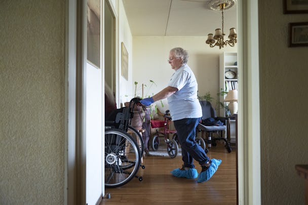 Marin Gabriel med blåa skoskydd leder en rullstol i ett rum med trägolv och diverse möbler och växter.