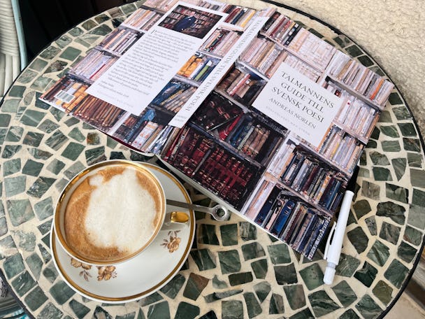 En kaffe i en vit och guldkantad kopp står på ett runt mosaikbord bredvid en uppslagen bok med titeln "Talmannens guide till svensk poesi" av Andreas Norlén.