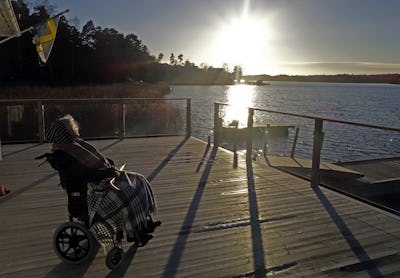 En äldre person i rullstol sitter på en träveranda med utsikt över en solbelyst sjö och träd i bakgrunden.