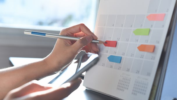 En person använder en penna för att markera ett datum på en kalender och håller samtidigt i en smartphone. Färgglada flikar är fästa vid olika datum på kalendern.