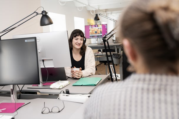 Bild av två kvinnor i ett kontor, en sitter vid ett skrivbord med två skärmar och verkar interagera med andra som står framför henne med ryggen mot kameran.