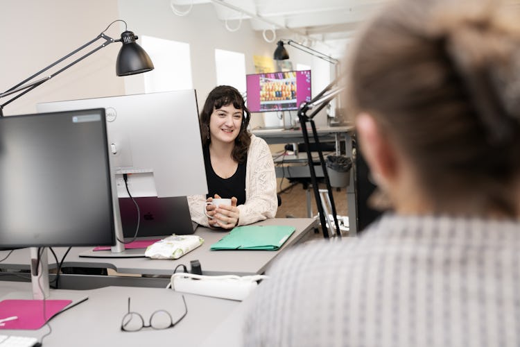 Bild av två kvinnor i ett kontor, en sitter vid ett skrivbord med två skärmar och verkar interagera med andra som står framför henne med ryggen mot kameran.