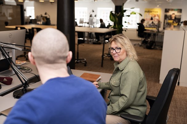 En kvinna som bär glasögon ses vid ett skrivbord i samtal med en skallig man klädd i blå skjorta, omgiven av aktiviteten i en ockuperad kontorsmiljö.
